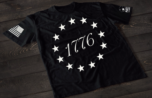 A black short-sleeve T-shirt laid flat on a dark wooden surface, featuring a circular arrangement of thirteen white stars surrounding the number “1776” in the center, inspired by the Betsy Ross flag. An American flag patch is displayed on one sleeve, with a small emblem printed on the other sleeve.