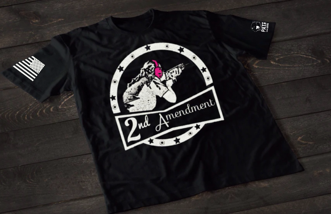 A black short-sleeve T-shirt laid flat on a dark wooden surface, featuring a white circular graphic of a woman aiming a rifle while wearing bright pink ear protection. The design includes stars around the circle and a banner reading “2nd Amendment.” An American flag patch is visible on one sleeve, and a small emblem is printed on the other sleeve.