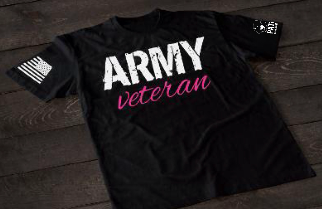 Black t-shirt on a wooden surface with bold white “ARMY” text and pink “veteran” script underneath. One sleeve shows an American flag, and the other has a small logo patch.