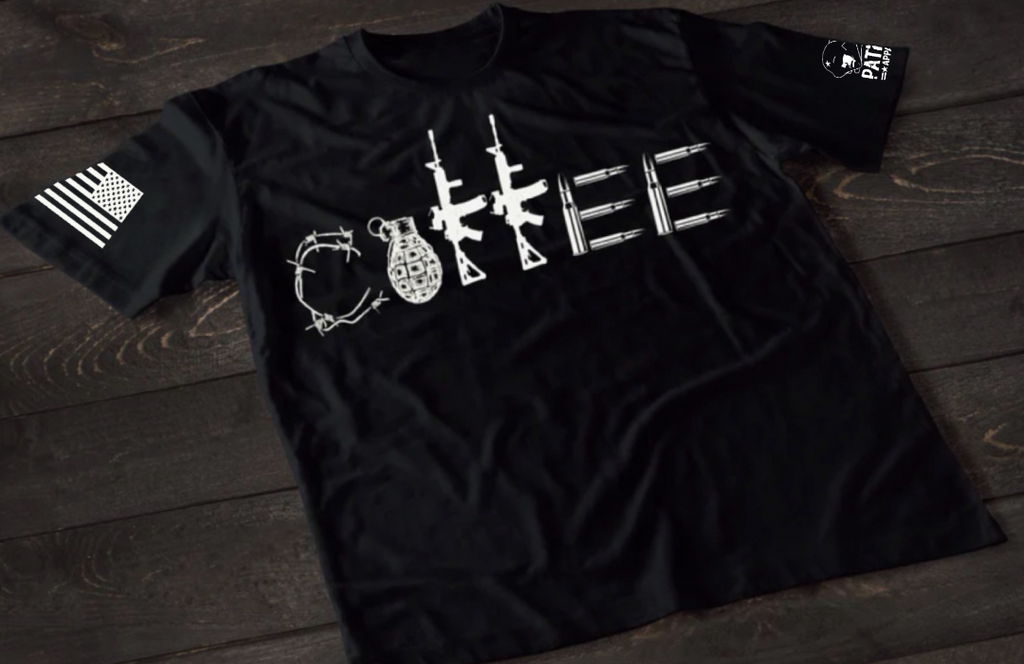 A black short-sleeve T-shirt laid flat on a dark wooden surface, featuring a stylized white graphic spelling “COFFEE,” with each letter formed by different tactical elements such as barbed wire, a grenade, rifles, and ammunition. An American flag patch is visible on one sleeve, and a small emblem is printed on the other sleeve.