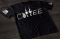 A black short-sleeve T-shirt laid flat on a dark wooden surface, featuring a stylized white graphic spelling “COFFEE,” with each letter formed by different tactical elements such as barbed wire, a grenade, rifles, and ammunition. An American flag patch is visible on one sleeve, and a small emblem is printed on the other sleeve.