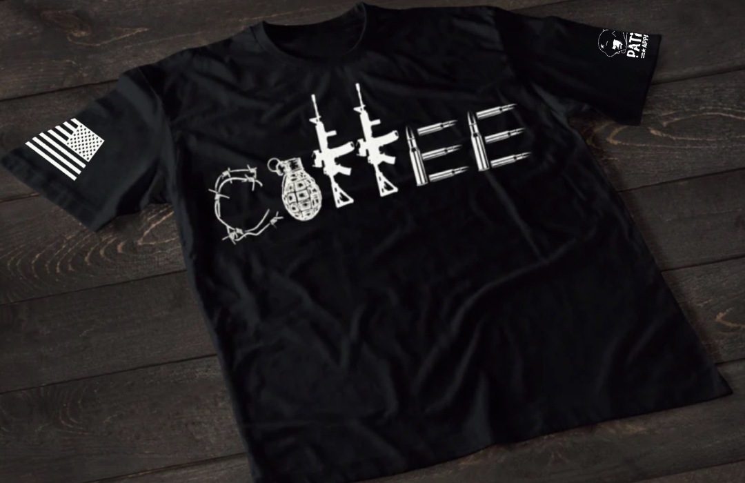 A black short-sleeve T-shirt laid flat on a dark wooden surface, featuring a stylized white graphic spelling “COFFEE,” with each letter formed by different tactical elements such as barbed wire, a grenade, rifles, and ammunition. An American flag patch is visible on one sleeve, and a small emblem is printed on the other sleeve.