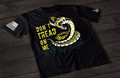 A black short-sleeve T-shirt laid flat on a dark wooden surface, featuring a bold graphic of a coiled rattlesnake in yellow and white with its mouth open. Beside the snake is the text “DON’T TREAD ON ME” in distressed yellow lettering. An American flag patch appears on one sleeve, and a small emblem is printed on the other sleeve.