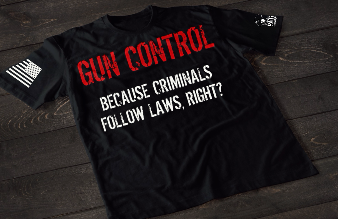 A black short-sleeve T-shirt laid flat on a dark wooden surface, featuring bold distressed text that reads “GUN CONTROL” in red, followed by “BECAUSE CRIMINALS FOLLOW LAWS, RIGHT?” in white. An American flag patch is visible on one sleeve, and a small emblem is printed on the other sleeve.