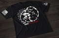 A black short-sleeve T-shirt laid flat on a dark wooden surface, featuring a white graphic of a battlefield memorial with a helmet, rifle, and boots alongside a silhouetted figure, surrounded by barbed wire and stars. The text reads “THIS SHIRT HONORS FALLEN HEROES,” with “FALLEN HEROES” highlighted in red. An American flag patch appears on one sleeve, and a small emblem is printed on the other sleeve.