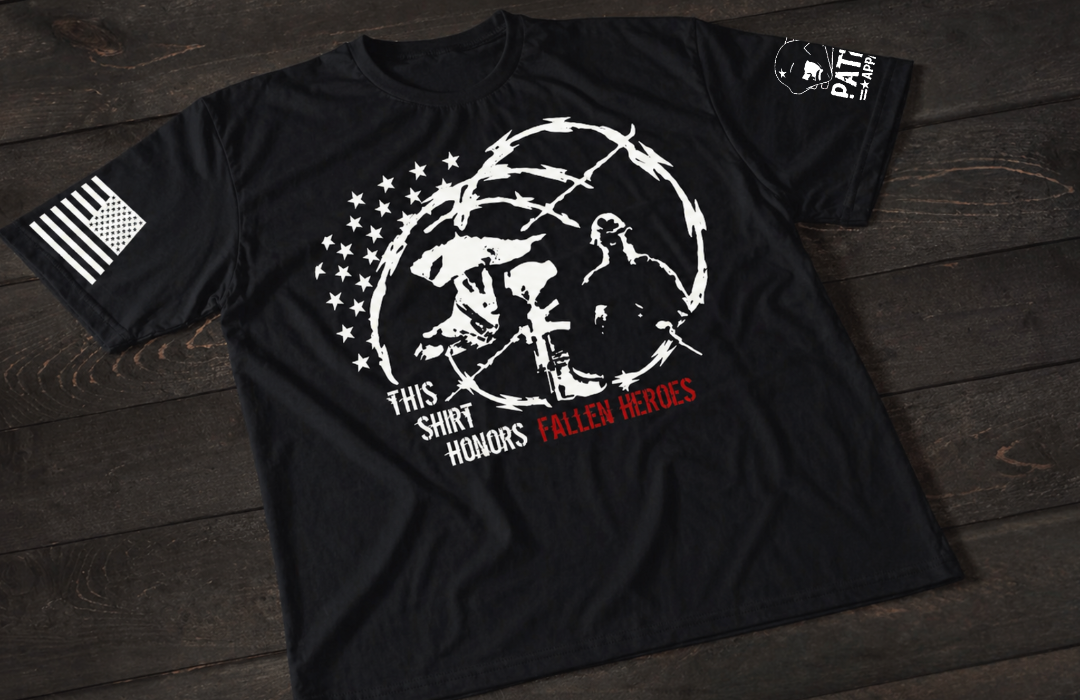A black short-sleeve T-shirt laid flat on a dark wooden surface, featuring a white graphic of a battlefield memorial with a helmet, rifle, and boots alongside a silhouetted figure, surrounded by barbed wire and stars. The text reads “THIS SHIRT HONORS FALLEN HEROES,” with “FALLEN HEROES” highlighted in red. An American flag patch appears on one sleeve, and a small emblem is printed on the other sleeve.