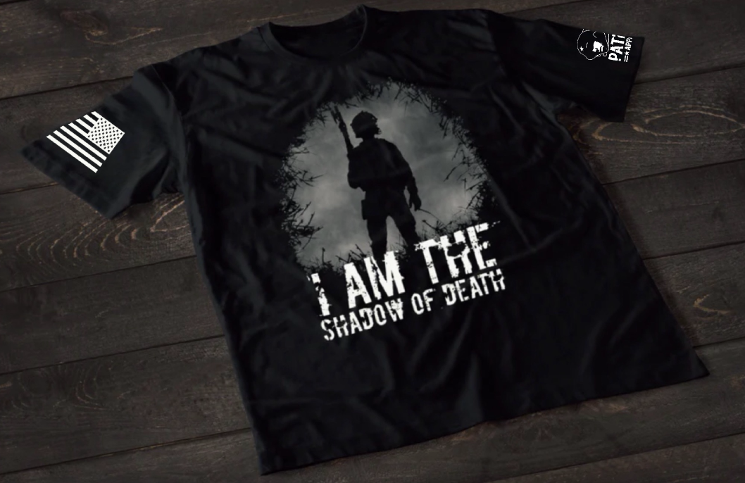 A black short-sleeve T-shirt laid flat on a dark wooden surface, featuring a grayscale graphic of a soldier silhouette holding a rifle, standing within a circular, shadowed scene. Below the image is the distressed text “I AM THE SHADOW OF DEATH.” An American flag patch appears on one sleeve, and a small emblem is printed on the other sleeve.