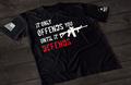 A black short-sleeve T-shirt laid flat on a dark wooden surface, featuring bold distressed text that reads “IT ONLY OFFENDS YOU UNTIL IT DEFENDS,” with the word “DEFENDS” in red. A white rifle graphic is incorporated into the design. An American flag patch is visible on one sleeve, and a small emblem is printed on the other sleeve.
