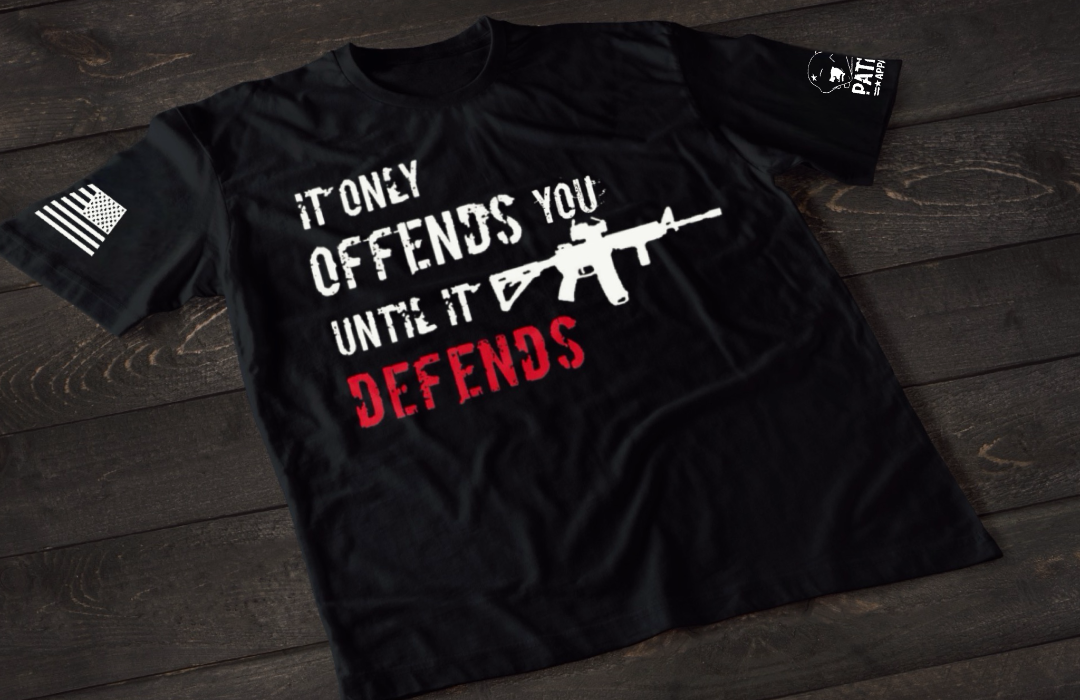 A black short-sleeve T-shirt laid flat on a dark wooden surface, featuring bold distressed text that reads “IT ONLY OFFENDS YOU UNTIL IT DEFENDS,” with the word “DEFENDS” in red. A white rifle graphic is incorporated into the design. An American flag patch is visible on one sleeve, and a small emblem is printed on the other sleeve.