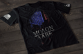 A black short-sleeve T-shirt laid flat on a dark wooden surface, featuring a stylized skull graphic filled with a distressed American flag pattern in blue and red tones. Below the image is the text “MOLON LABE” with a spear running through the lettering. An American flag patch is visible on one sleeve, and a small emblem is printed on the other sleeve.