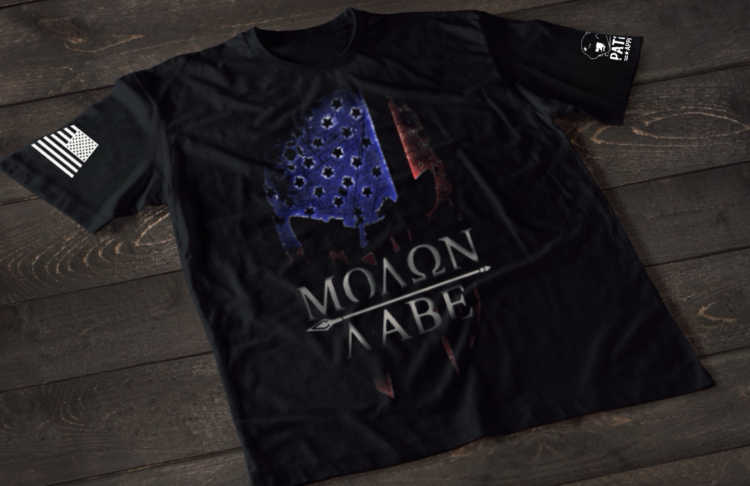 A black short-sleeve T-shirt laid flat on a dark wooden surface, featuring a stylized skull graphic filled with a distressed American flag pattern in blue and red tones. Below the image is the text “MOLON LABE” with a spear running through the lettering. An American flag patch is visible on one sleeve, and a small emblem is printed on the other sleeve.