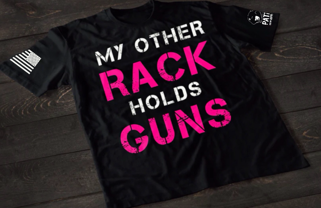 A black short-sleeve T-shirt laid flat on a dark wooden surface, featuring large distressed text that reads “MY OTHER RACK HOLDS GUNS,” with “RACK” and “GUNS” in bright pink and the rest in white. An American flag patch is visible on one sleeve, and a small emblem is printed on the other sleeve.