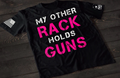 A black short-sleeve T-shirt laid flat on a dark wooden surface, featuring large distressed text that reads “MY OTHER RACK HOLDS GUNS,” with “RACK” and “GUNS” in bright pink and the rest in white. An American flag patch is visible on one sleeve, and a small emblem is printed on the other sleeve.