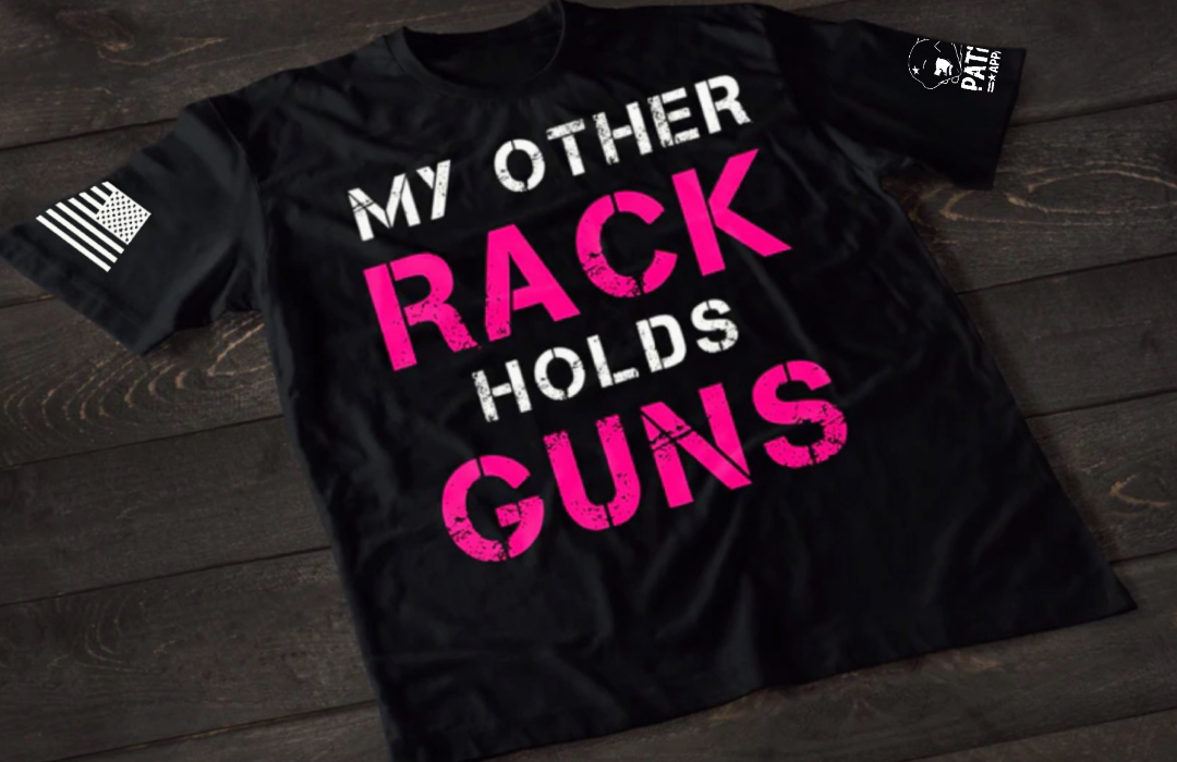 A black short-sleeve T-shirt laid flat on a dark wooden surface, featuring large distressed text that reads “MY OTHER RACK HOLDS GUNS,” with “RACK” and “GUNS” in bright pink and the rest in white. An American flag patch is visible on one sleeve, and a small emblem is printed on the other sleeve.