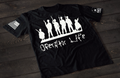 A black short-sleeve T-shirt laid flat on a dark wooden surface, featuring a white graphic of multiple soldier silhouettes holding rifles in different stances. Below the figures is the stylized text “Operator Life.” An American flag patch appears on one sleeve, and a small emblem is printed on the other sleeve.