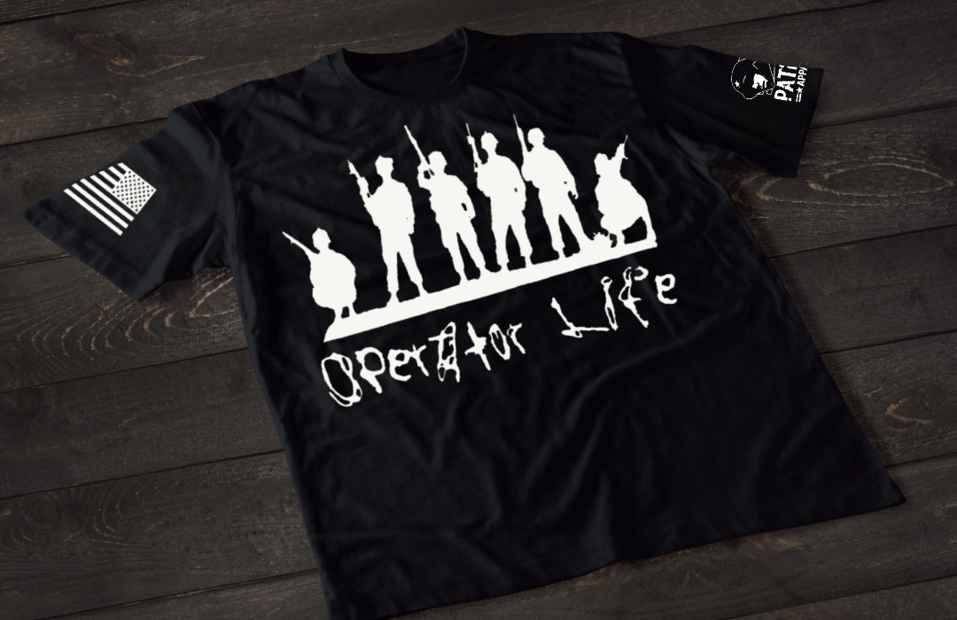 A black short-sleeve T-shirt laid flat on a dark wooden surface, featuring a white graphic of multiple soldier silhouettes holding rifles in different stances. Below the figures is the stylized text “Operator Life.” An American flag patch appears on one sleeve, and a small emblem is printed on the other sleeve.
