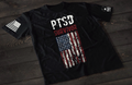 A black short-sleeve T-shirt laid flat on a dark wooden surface, featuring the text “PTSD SURVIVOR” in distressed white and red lettering above a worn, vertical American flag graphic. An American flag patch is visible on one sleeve, and a small emblem is printed on the other sleeve.