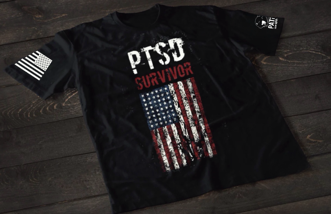 A black short-sleeve T-shirt laid flat on a dark wooden surface, featuring the text “PTSD SURVIVOR” in distressed white and red lettering above a worn, vertical American flag graphic. An American flag patch is visible on one sleeve, and a small emblem is printed on the other sleeve.