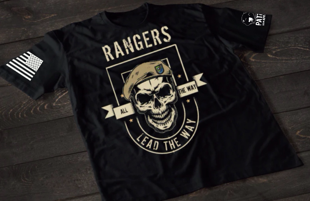 A black short-sleeve T-shirt laid flat on a dark wooden surface, featuring a large central graphic of a skull wearing a military beret inside a shield design. The text “RANGERS” appears at the top, with “ALL THE WAY” and “LEAD THE WAY” incorporated into the design. An American flag patch is visible on one sleeve, and a small emblem is printed on the other sleeve.