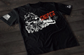 A black short-sleeve T-shirt laid flat on a dark wooden surface, featuring a distressed white graphic of a sniper rifle set against a rough, textured background. The word “SNIPER” appears above in bold red lettering, with a small crosshair symbol to the side. An American flag patch is visible on one sleeve, and a small emblem is printed on the other sleeve.