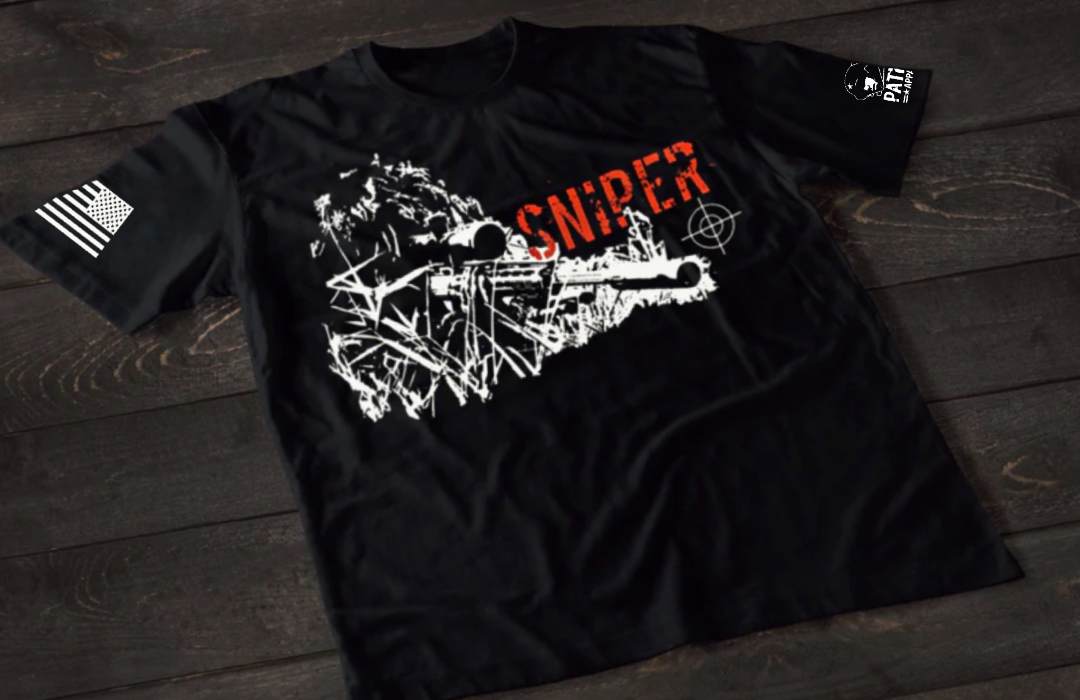 A black short-sleeve T-shirt laid flat on a dark wooden surface, featuring a distressed white graphic of a sniper rifle set against a rough, textured background. The word “SNIPER” appears above in bold red lettering, with a small crosshair symbol to the side. An American flag patch is visible on one sleeve, and a small emblem is printed on the other sleeve.