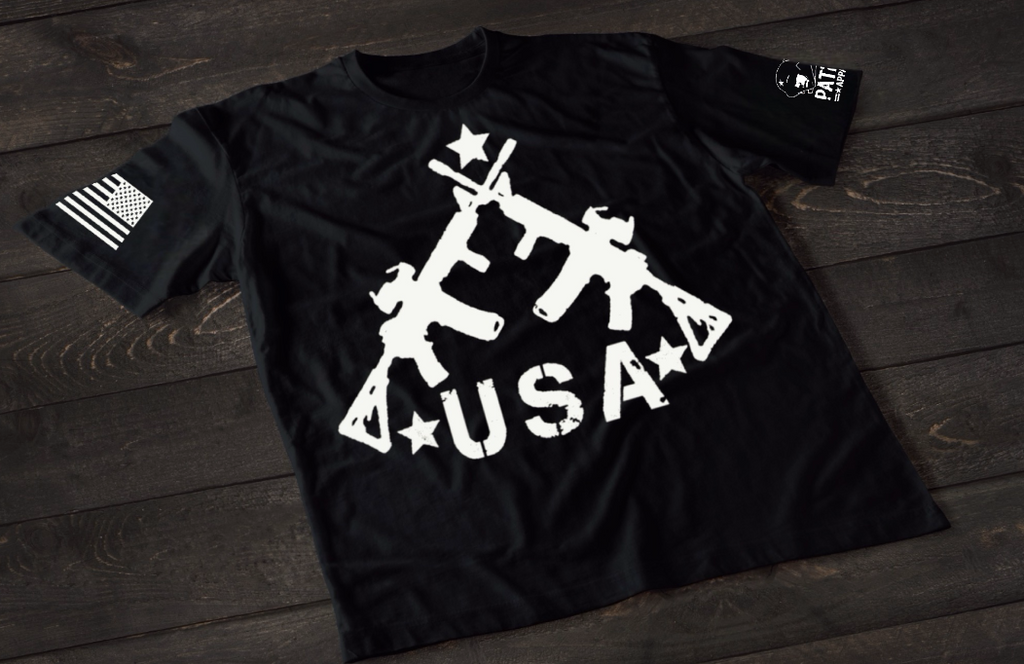 A black short-sleeve T-shirt laid flat on a dark wooden surface, featuring a bold white graphic of three crossed rifles above the word “USA” with stars. An American flag patch appears on one sleeve, and a small circular emblem is printed on the other sleeve.