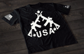 A black short-sleeve T-shirt laid flat on a dark wooden surface, featuring a bold white graphic of three crossed rifles above the word “USA” with stars. An American flag patch appears on one sleeve, and a small circular emblem is printed on the other sleeve.