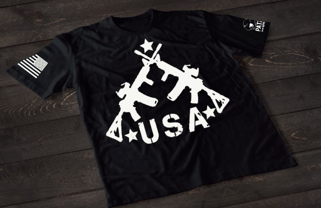 A black short-sleeve T-shirt laid flat on a dark wooden surface, featuring a bold white graphic of three crossed rifles above the word “USA” with stars. An American flag patch appears on one sleeve, and a small circular emblem is printed on the other sleeve.