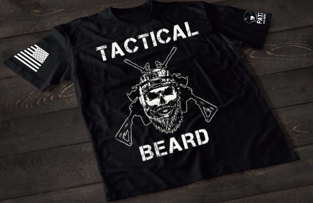 A black short-sleeve T-shirt laid flat on a dark wooden surface, featuring a bold white graphic of a bearded skull wearing tactical headgear with crossed rifles behind it. The word “TACTICAL” is printed above the design and “BEARD” below. An American flag patch is visible on one sleeve, and a small emblem is printed on the other sleeve.