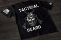 A black short-sleeve T-shirt laid flat on a dark wooden surface, featuring a bold white graphic of a bearded skull wearing tactical headgear with crossed rifles behind it. The word “TACTICAL” is printed above the design and “BEARD” below. An American flag patch is visible on one sleeve, and a small emblem is printed on the other sleeve.