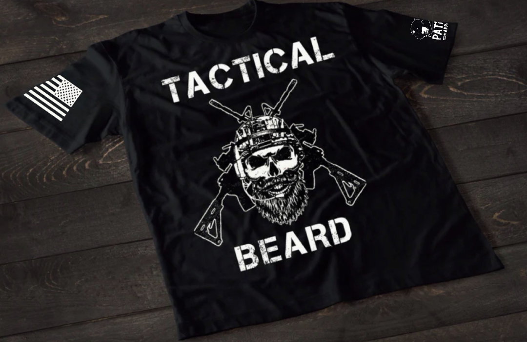 A black short-sleeve T-shirt laid flat on a dark wooden surface, featuring a bold white graphic of a bearded skull wearing tactical headgear with crossed rifles behind it. The word “TACTICAL” is printed above the design and “BEARD” below. An American flag patch is visible on one sleeve, and a small emblem is printed on the other sleeve.