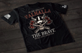 A black short-sleeve T-shirt laid flat on a dark wooden surface, featuring a detailed graphic of a skull wearing a Viking helmet with crossed axes behind it. The text reads “IN THE HALLS OF VALHALLA” above and “THE BRAVE SHALL LIVE FOREVER” below. An American flag patch appears on one sleeve, and a small emblem is printed on the other sleeve.