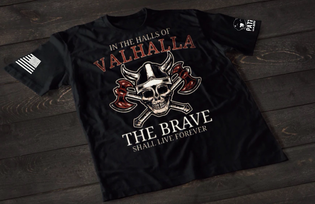 A black short-sleeve T-shirt laid flat on a dark wooden surface, featuring a detailed graphic of a skull wearing a Viking helmet with crossed axes behind it. The text reads “IN THE HALLS OF VALHALLA” above and “THE BRAVE SHALL LIVE FOREVER” below. An American flag patch appears on one sleeve, and a small emblem is printed on the other sleeve.