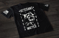 A black short-sleeve T-shirt laid flat on a dark wooden surface, featuring a distressed white graphic of a soldier with hands clasped in front of their face. Surrounding the image is the text “VETERANS LIVES MATTER” arranged in a bold, worn-style font with stars. An American flag patch is visible on one sleeve, and a small emblem is printed on the other sleeve.