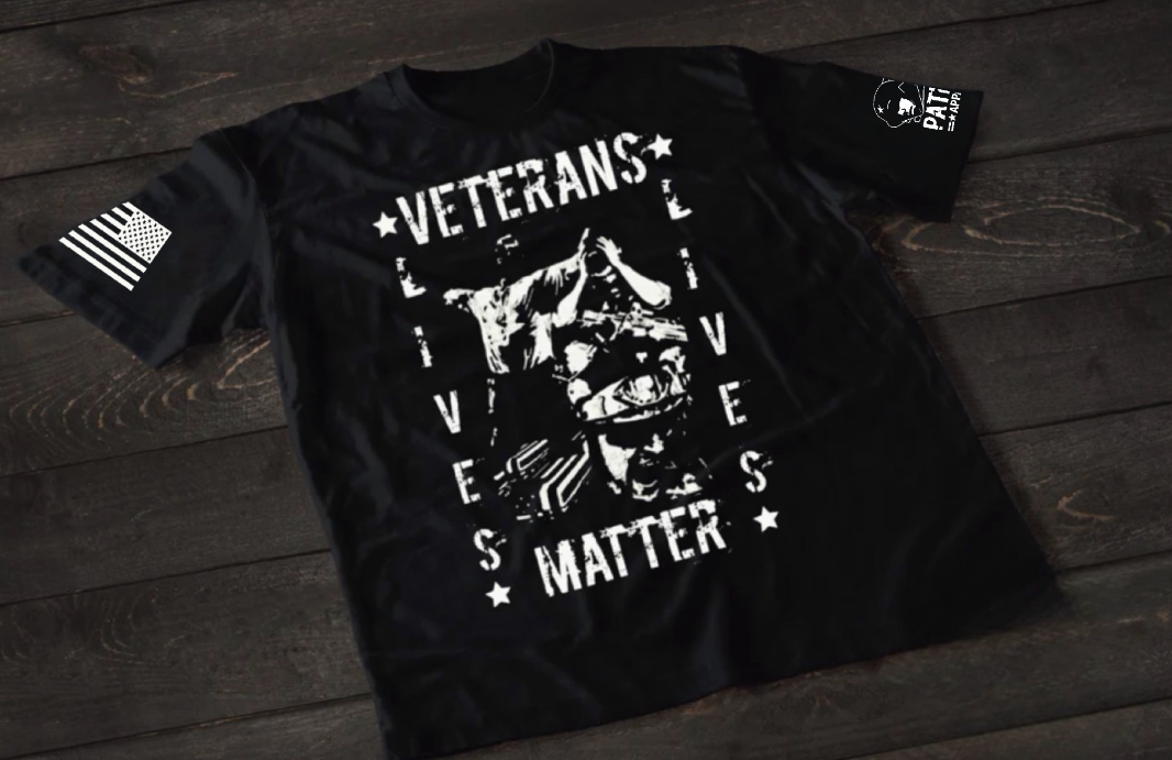 A black short-sleeve T-shirt laid flat on a dark wooden surface, featuring a distressed white graphic of a soldier with hands clasped in front of their face. Surrounding the image is the text “VETERANS LIVES MATTER” arranged in a bold, worn-style font with stars. An American flag patch is visible on one sleeve, and a small emblem is printed on the other sleeve.