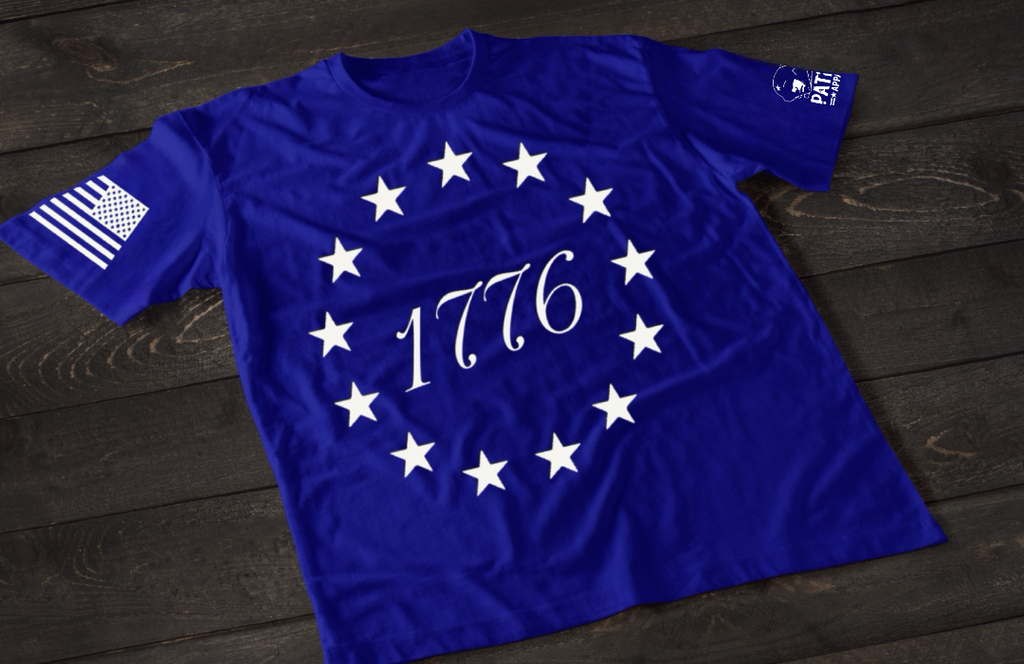 A blue short-sleeve T-shirt laid flat on a dark wooden surface, featuring a circular arrangement of thirteen white stars surrounding the number “1776” in the center, inspired by the Betsy Ross flag. An American flag patch is displayed on one sleeve, with a small emblem printed on the other sleeve.
