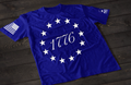 A blue short-sleeve T-shirt laid flat on a dark wooden surface, featuring a circular arrangement of thirteen white stars surrounding the number “1776” in the center, inspired by the Betsy Ross flag. An American flag patch is displayed on one sleeve, with a small emblem printed on the other sleeve.