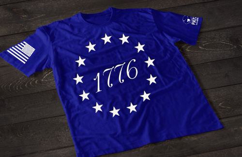 A blue short-sleeve T-shirt laid flat on a dark wooden surface, featuring a circular arrangement of thirteen white stars surrounding the number “1776” in the center, inspired by the Betsy Ross flag. An American flag patch is displayed on one sleeve, with a small emblem printed on the other sleeve.