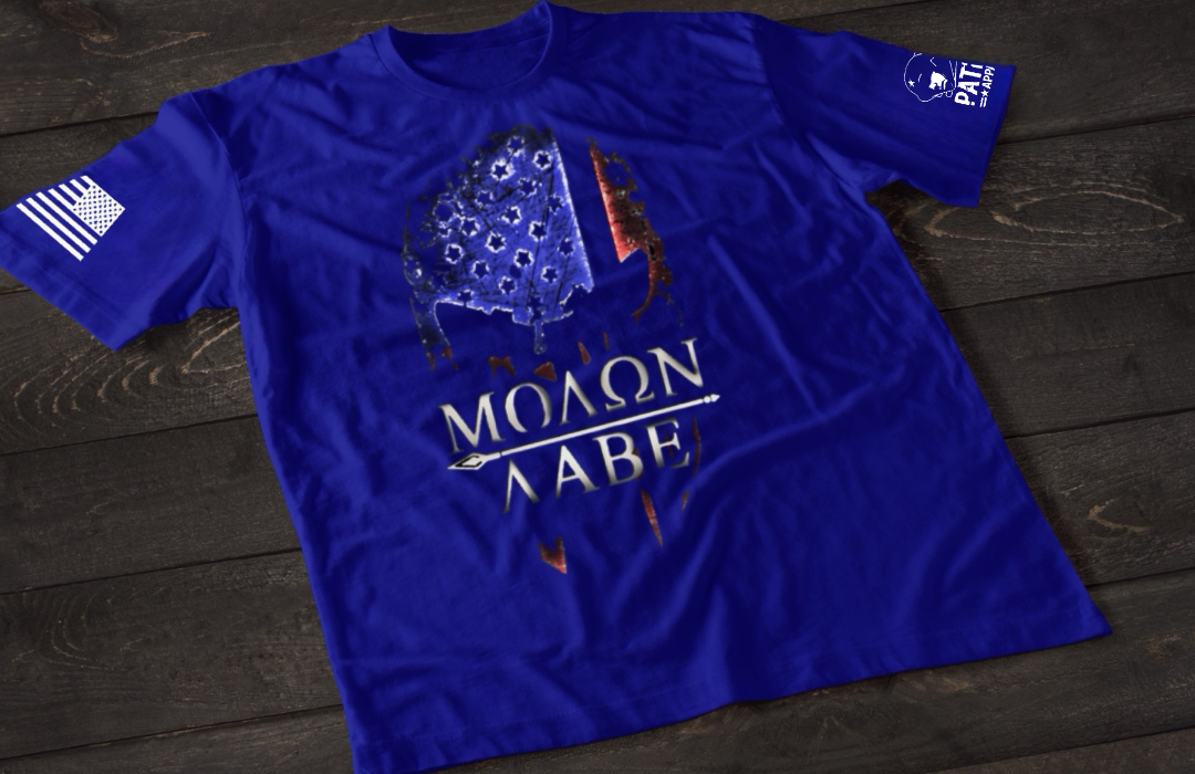 A blue short-sleeve T-shirt laid flat on a dark wooden surface, featuring a stylized skull graphic filled with a distressed American flag pattern in blue and red tones. Below the image is the text “MOLON LABE” with a spear running through the lettering. An American flag patch is visible on one sleeve, and a small emblem is printed on the other sleeve.