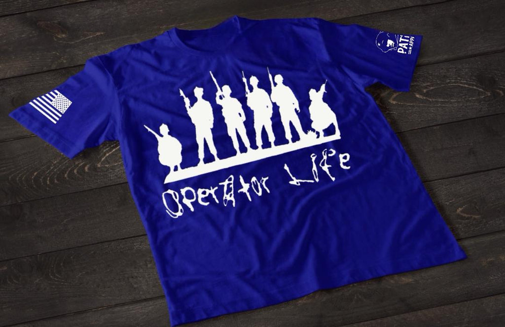 A blue short-sleeve T-shirt laid flat on a dark wooden surface, featuring a white graphic of multiple soldier silhouettes holding rifles in different stances. Below the figures is the stylized text “Operator Life.” An American flag patch appears on one sleeve, and a small emblem is printed on the other sleeve.