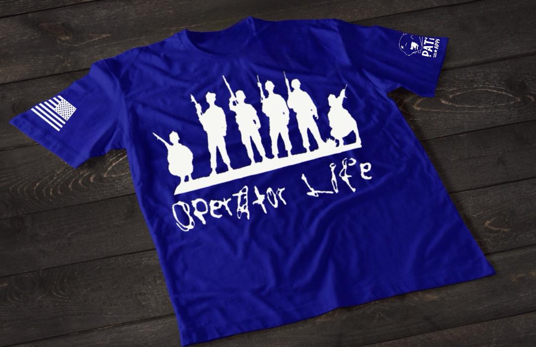 A blue short-sleeve T-shirt laid flat on a dark wooden surface, featuring a white graphic of multiple soldier silhouettes holding rifles in different stances. Below the figures is the stylized text “Operator Life.” An American flag patch appears on one sleeve, and a small emblem is printed on the other sleeve.
