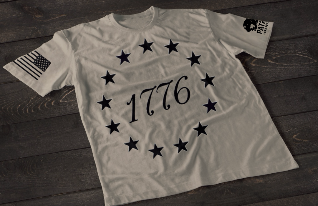 A gray short-sleeve T-shirt laid flat on a dark wooden surface, featuring a circular arrangement of thirteen white stars surrounding the number “1776” in the center, inspired by the Betsy Ross flag. An American flag patch is displayed on one sleeve, with a small emblem printed on the other sleeve.