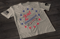 A gray short-sleeve T-shirt laid flat on a dark wooden surface, featuring a circular arrangement of red and blue stars surrounding the text “2nd Amendment” in bold lettering, with “est. 1791” displayed on a ribbon below. An American flag patch appears on one sleeve, and a small emblem is printed on the other sleeve.