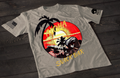 A gray short-sleeve T-shirt laid flat on a dark wooden surface, featuring a colorful circular graphic of a tropical beach scene with palm trees, ocean waves, and a sunset in red and yellow tones. The word “ALOHA” appears above the design, with “SNACK BAR” printed below in a stylized font. An American flag patch is visible on one sleeve, and a small emblem is printed on the other sleeve.