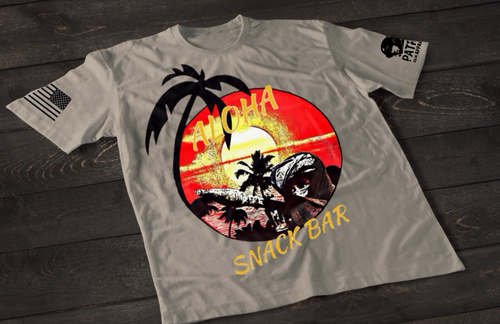 A gray short-sleeve T-shirt laid flat on a dark wooden surface, featuring a colorful circular graphic of a tropical beach scene with palm trees, ocean waves, and a sunset in red and yellow tones. The word “ALOHA” appears above the design, with “SNACK BAR” printed below in a stylized font. An American flag patch is visible on one sleeve, and a small emblem is printed on the other sleeve.