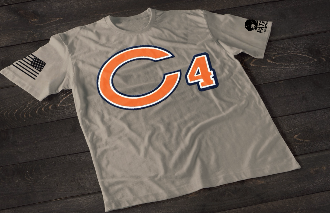 A gray short-sleeve T-shirt laid flat on a dark wooden surface, featuring a large orange letter “C” outlined in navy blue with the number “4” beside it in matching colors. An American flag patch is visible on one sleeve, and a small emblem is printed on the other sleeve.