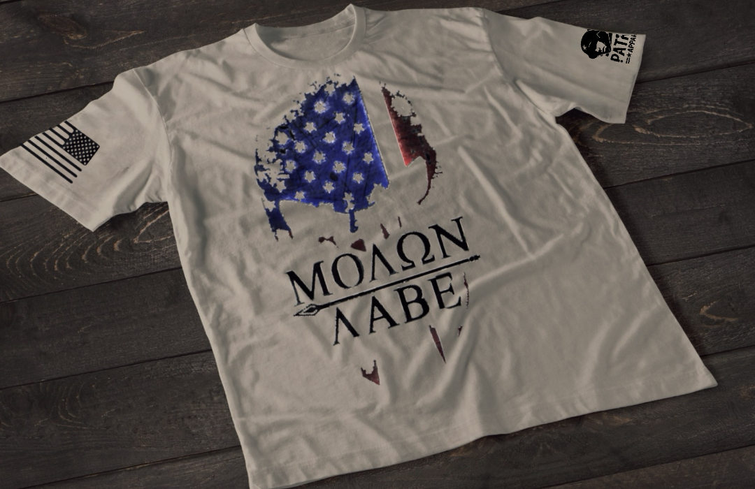 A gray short-sleeve T-shirt laid flat on a dark wooden surface, featuring a stylized skull graphic filled with a distressed American flag pattern in blue and red tones. Below the image is the text “MOLON LABE” with a spear running through the lettering. An American flag patch is visible on one sleeve, and a small emblem is printed on the other sleeve.