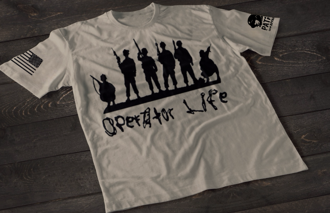 A gray short-sleeve T-shirt laid flat on a dark wooden surface, featuring a white graphic of multiple soldier silhouettes holding rifles in different stances. Below the figures is the stylized text “Operator Life.” An American flag patch appears on one sleeve, and a small emblem is printed on the other sleeve.