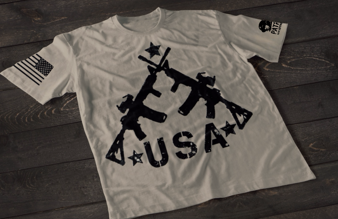 A gray short-sleeve T-shirt laid flat on a dark wooden surface, featuring a bold white graphic of three crossed rifles above the word “USA” with stars. An American flag patch appears on one sleeve, and a small circular emblem is printed on the other sleeve.
