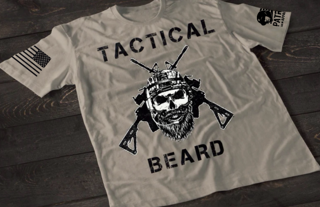 A gray short-sleeve T-shirt laid flat on a dark wooden surface, featuring a bold white graphic of a bearded skull wearing tactical headgear with crossed rifles behind it. The word “TACTICAL” is printed above the design and “BEARD” below. An American flag patch is visible on one sleeve, and a small emblem is printed on the other sleeve.